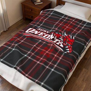 May include: A red, black, and white plaid blanket with the text "SUNY ONEONTA" in white and red, and a red dragon graphic. The blanket is folded over a bed.