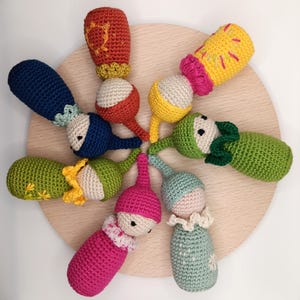 Puede incluir: Una colección de muñecas de ganchillo hechas a mano en varios colores, incluyendo rojo, azul, verde, amarillo y rosa. Cada muñeca tiene un diseño único, algunas con detalles bordados y cuellos decorativos. Las muñecas están dispuestas sobre una superficie de madera clara.
