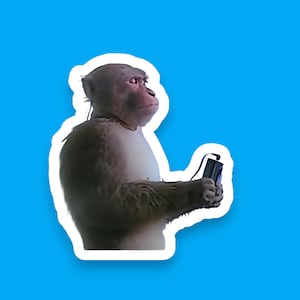 May include: A sticker featuring a monkey holding a small, black electronic device with a cord. The monkey is light brown with a white outline, set against a bright blue background.