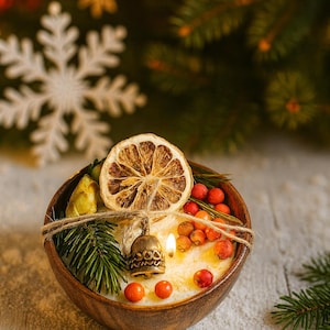 May include: A wooden bowl candle with a dried orange slice, berries, and a small bell. The candle is decorated with a sprig of evergreen and tied with twine. The background features a snowflake and Christmas tree branches.