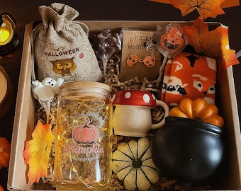 May include: A Halloween-themed gift box filled with festive items. Includes a jar with a pumpkin graphic, a mushroom mug, a black cauldron, and a bag with an owl. The box also contains a pair of orange socks, a pumpkin-shaped candy, and decorative leaves.
