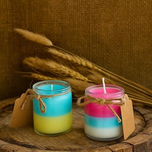 May include: Two small, layered candles in glass jars. One candle has blue and yellow layers, the other has pink, blue, and white layers. Each jar is tied with twine and has a blank tag. The candles sit on a wooden surface.