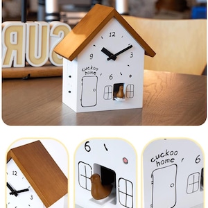 May include: A white cuckoo clock shaped like a house with a brown roof. The clock has black hands, a door, windows, and the words "cuckoo home." A small brown bird is visible in the window. The clock is on a wooden surface.