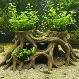 May include: A brown resin aquarium ornament shaped like a tree stump with exposed roots and two hollow tops containing bright green aquatic plants. The structure features multiple arched openings and sits on a light-coloured sandy substrate.