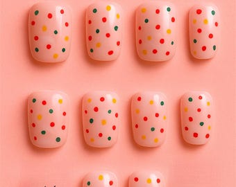 Peut inclure: Dix faux ongles roses ornés de pois rouges, jaunes et verts. Le texte "DOTTY & CUTE" est affiché au-dessus des ongles, avec le logo "Muse Nail Arts" en bas. L'arrière-plan est rose clair.