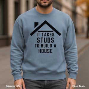May include: Blue jean crewneck sweatshirt with the text "IT TAKES STUDS TO BUILD A HOUSE" in black. The graphic includes a black house outline above the text. The sweatshirt is a solid blue color.