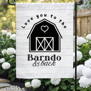 May include: A white garden flag with a black barn illustration and the text "Love you to the Barndo & back." The flag is hanging on a black metal stand, surrounded by white flowers and greenery.
