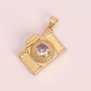 Peut inclure: Pendentif doré en forme d'appareil photo avec une gemme ronde et transparente au centre. Le pendentif présente un design détaillé, avec un objectif et un corps rectangulaire. La bélière est également dorée.