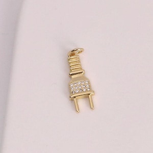 May include: A gold-colored pendant in the shape of an electrical plug. The plug has a rectangular body with a section covered in small, clear crystals. The prongs are visible at the bottom. The pendant is on a white surface.