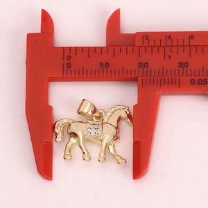 Könnte beinhalten: Ein goldfarbener Pferdeanhänger mit klaren Strasssteinen. Der Anhänger wird mit einer Schieblehre gemessen und ist etwa 2,5 cm lang.