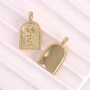 Peut inclure: Deux pendentifs dorés. L'un présente un motif détaillé d'un palmier et d'un serpent dans un cadre en forme d'arche. L'autre pendentif a une surface lisse et vierge. Les deux ont une petite boucle en haut pour une chaîne.