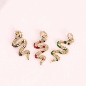 Pode incluir: Três pingentes de cobra dourados com olhos pretos e uma argola no topo para pendurar. Uma cobra tem detalhes vermelhos e dourados, outra tem detalhes verdes e dourados, e a terceira tem detalhes pretos e dourados.