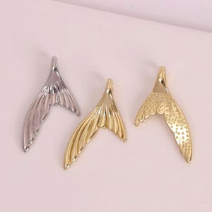 Puede incluir: Tres colgantes de cola de sirena en plata, oro y oro texturizado. Estos colgantes metálicos tienen un lazo para sujetarlos a joyas. Las colas están desplegadas y detalladas con líneas y texturas.