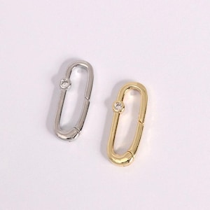 May include: Two oval-shaped clasps, one silver-toned and one gold-toned, each with a small, clear gemstone. The clasps are displayed on a white background. These jewelry findings are suitable for necklaces, bracelets, or other accessories.