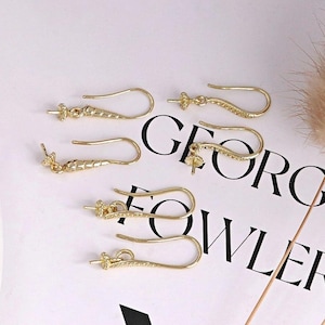 May include: Several gold-colored earring hooks are arranged on a white surface. The hooks have a curved design with a small decorative element at the base. The hooks are designed for jewelry making.