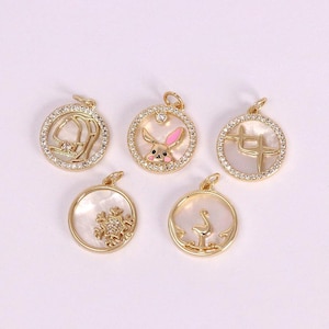 May include: Five gold-colored pendants with various designs. Each pendant has a round shape with a white mother-of-pearl background. The designs include a bunny, a snowflake, a swan, and other shapes, all surrounded by a ring of small, clear crystals.