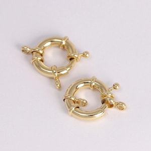 May include: Two gold-colored, round jewelry clasps. Each clasp has a circular body with a hinged opening and a small, decorative ball on the end. The clasps are designed to secure necklaces or bracelets.