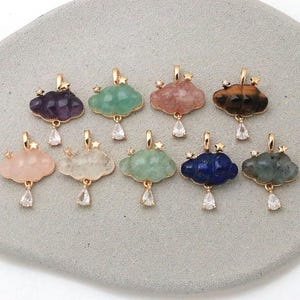 Könnte beinhalten: Eine Sammlung von zehn wolkenförmigen Anhängern in verschiedenen Farben wie Lila, Grün, Rosa, Braun und Blau. Jeder Anhänger verfügt über einen goldfarbenen Metallrahmen, einen kleinen Sternakzent und einen klaren, tropfenförmigen Kristallanhänger.