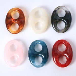 Könnte beinhalten: Sechs ovale Harzperlen in verschiedenen Farben. Jede Perle hat zwei runde Löcher. Die Farben sind Braun, Creme, Schwarz, Rosa, Blau und Rot. Diese Perlen eignen sich für die Schmuckherstellung oder Bastelprojekte.