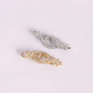 May include: Two decorative jewelry clasps, one silver-toned and one gold-toned, are displayed against a white background. Both clasps feature a series of small, sparkling stones and a unique interlocking design, perfect for necklaces or bracelets.