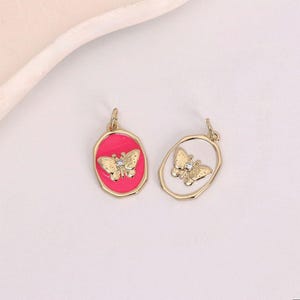 May include: Two gold-colored butterfly pendants. One pendant features a butterfly on a pink oval background, while the other has a butterfly within a gold frame. Both have a small crystal accent.