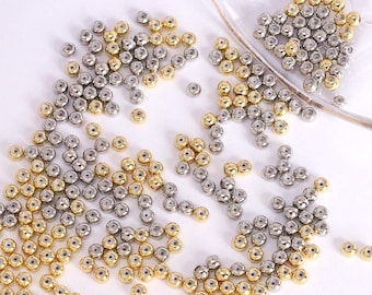 May include: A collection of small, round beads in gold and silver tones scattered across a white surface. The beads appear to be metallic and have a small hole through the center. Some beads are in a clear container.