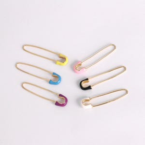 May include: Six gold-colored safety pins with colorful enamel accents. The pins feature yellow, pink, black, white, blue, and purple enamel on the curved end. The pins are arranged on a white surface.