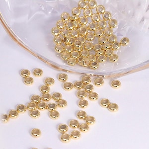 May include: A collection of small, shiny, gold-colored metal beads. The beads are round with a hole in the center, and are scattered on a white surface and inside a clear dish. These beads are likely used for jewelry making or other crafts.