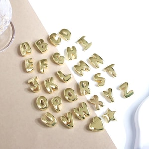 Könnte beinhalten: Eine Sammlung goldfarbener Buchstabenanhänger, einschließlich der Buchstaben A bis Z, eines Sterns und eines Herzens. Die Anhänger sind auf einer weißen und beigefarbenen Oberfläche mit glänzendem Finish angeordnet. Die Buchstaben sind in einer abgerundeten Schriftart.