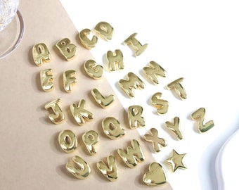 May include: A collection of gold-colored alphabet charms, including letters A through Z, a star, and a heart. The charms are arranged on a white and tan surface, with a glossy finish. The letters are in a rounded, bubble-style font.