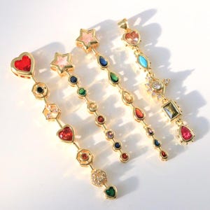 Puede incluir: Cuatro pendientes dorados con detalles de gemas de colores. Cada pendiente presenta una serie de gemas de varias formas, incluyendo corazones, estrellas y lágrimas. Las gemas son de color rojo, rosa, azul, verde y amarillo. Sobre una superficie blanca.
