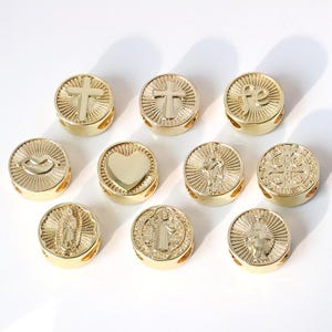 Puede incluir: Una colección de cuentas redondas doradas con varios diseños religiosos y simbólicos. Las cuentas presentan cruces, corazones y figuras, con una superficie brillante y reflectante. Las cuentas están dispuestas sobre una superficie blanca.