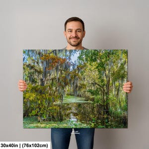 A man is holding a large framed print of a lush, forested landscape with a pond in the center.