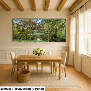 The image is a large, three-panel painting depicting a serene landscape with a pond surrounded by lush greenery. The painting is hung on a wall above a wooden dining table with four chairs. The table is set with a vase of white flowers, adding a touch of elegance to the scene. The overall composition and arrangement of the elements create a harmonious and inviting atmosphere in the room.