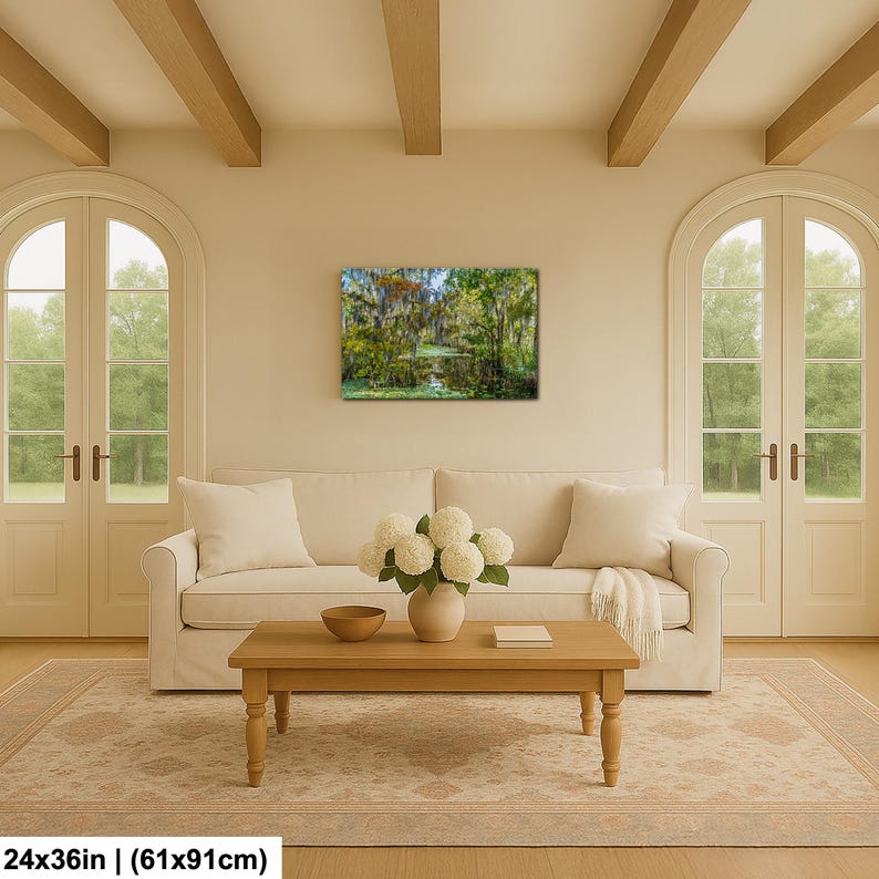 a cozy living room with a large window overlooking a lush green forest. The room is furnished with a white sofa, a wooden coffee table, and a vase of white flowers. The walls are painted in a light color, and the ceiling features wooden beams. A painting of a forest scene hangs on the wall, adding a touch of nature to the space.