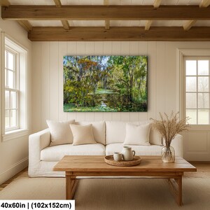 a cozy living room with a large, vibrant painting of a lush forest scene hanging on the wall above a white sofa. The painting features a tranquil pond surrounded by trees and foliage, creating a serene and natural atmosphere. The room is furnished with a wooden coffee table and a vase of dried flowers, adding a touch of elegance and warmth to the space.