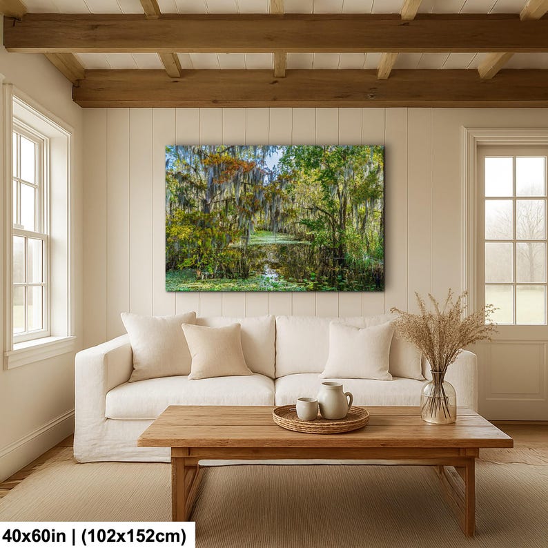 a cozy living room with a large, vibrant painting of a lush forest scene hanging on the wall above a white sofa. The painting features a tranquil pond surrounded by trees and foliage, creating a serene and natural atmosphere. The room is furnished with a wooden coffee table and a vase of dried flowers, adding a touch of elegance and warmth to the space.