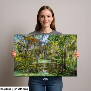 A woman is holding a large canvas print of a lush, forested landscape with a pond in the center.