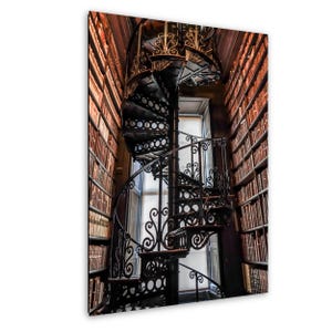 May include: A black wrought iron spiral staircase in a library. The staircase features decorative scrollwork and ascends. Bookshelves filled with books flank the staircase. A window is visible in the background, with natural light.