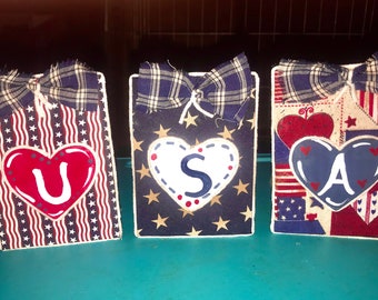 May include: Three wooden blocks with red, white, and blue fabric, each with a heart-shaped cutout containing a letter. The blocks spell out "USA".