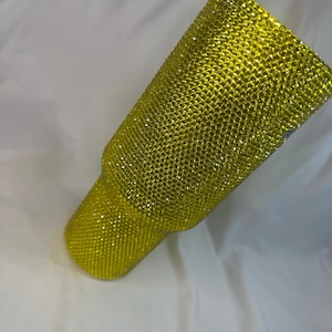 May include: A cylindrical tumbler fully covered in small, bright yellow rhinestones. The surface features a dense, uniform grid of sparkling, faceted stones that create a textured, reflective appearance across the entire exterior of the vessel.