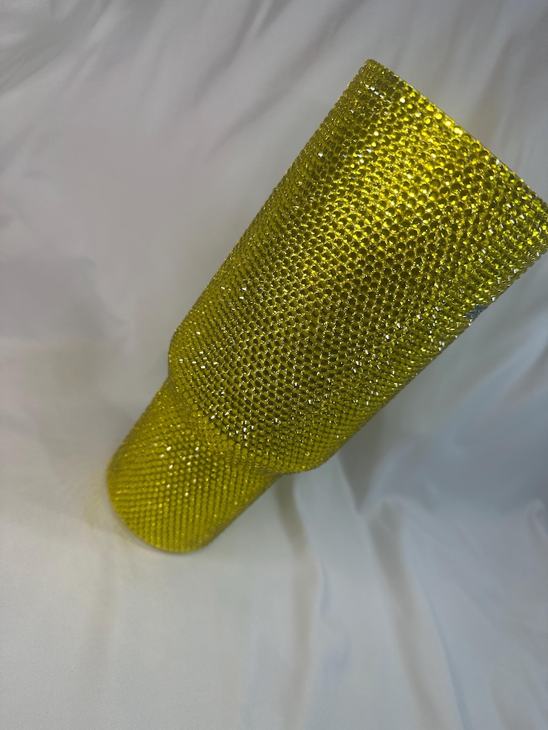 May include: A cylindrical tumbler fully covered in small, bright yellow rhinestones. The surface features a dense, uniform grid of sparkling, faceted stones that create a textured, reflective appearance across the entire exterior of the vessel.