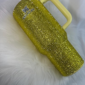 May include: A yellow tumbler covered in small, reflective rhinestones with a light yellow handle. The surface is fully encrusted with sparkling yellow crystals, and the word Stanley is visible in silver rhinestones near the top of the cup.