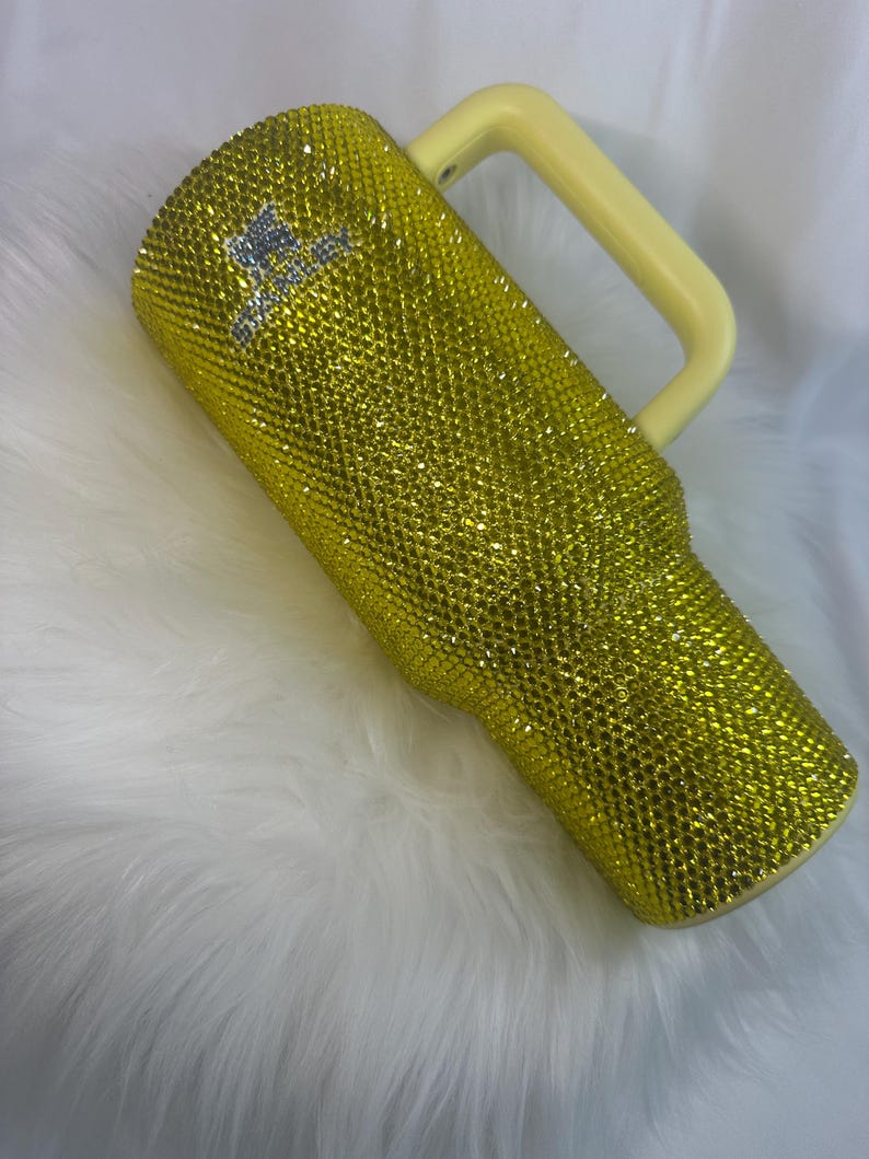 May include: A yellow tumbler covered in small, reflective rhinestones with a light yellow handle. The surface is fully encrusted with sparkling yellow crystals, and the word Stanley is visible in silver rhinestones near the top of the cup.