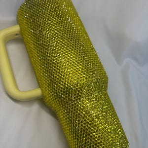 May include: A tall, cylindrical tumbler with a handle, fully covered in small, reflective yellow rhinestones. The tumbler features a light yellow plastic handle and a matching light yellow lid, creating a uniform, bright, and textured appearance.