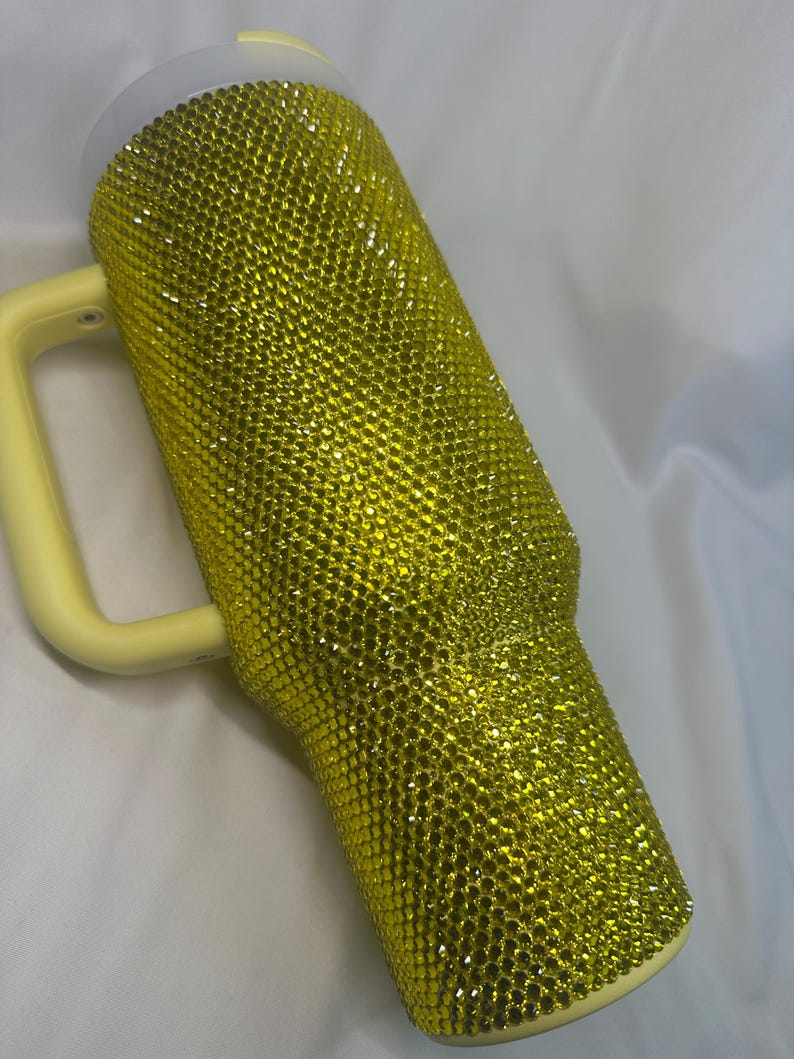 May include: A tall, cylindrical tumbler with a handle, fully covered in small, reflective yellow rhinestones. The tumbler features a light yellow plastic handle and a matching light yellow lid, creating a uniform, bright, and textured appearance.