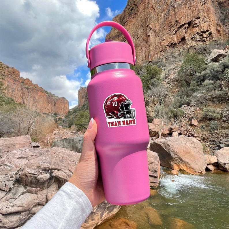 May include: A matte pink insulated water bottle with a handle on the lid. The bottle features a sticker of a dark red football helmet with the number 10, the text ANYNAME, and the text TEAM NAME printed in white on the front of the helmet graphic.