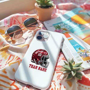 May include: A white smartphone case featuring a sticker of a maroon football helmet with the number 10, the text ANYNAME above it, and the text TEAM NAME below it. The case has a triple-lens camera cutout on the upper left corner.