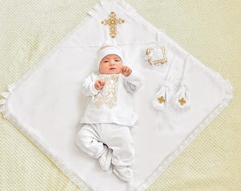 Puede incluir: Un bebé con un conjunto blanco con bordados dorados, un gorro a juego y patucos, tumbado sobre una manta blanca con una cruz dorada y ribete de encaje. La manta tiene un babero a juego y dos bolsitas.