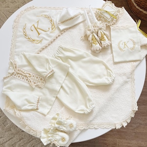 Puede incluir: Un conjunto de bautizo para bebé de color crema, que incluye una manta, un gorro, una camisa, un pantalón, patucos y accesorios. El conjunto presenta bordados dorados, ribetes de encaje y un diseño con la letra "K" monogramada.